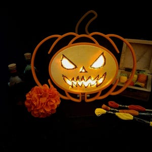 May include: An orange plastic pumpkin frame with a cross-stitch mesh center featuring a glowing jack-o'-lantern face. The design includes sharp, illuminated eyes and a jagged mouth. Nearby are an orange fabric flower and embroidery floss.