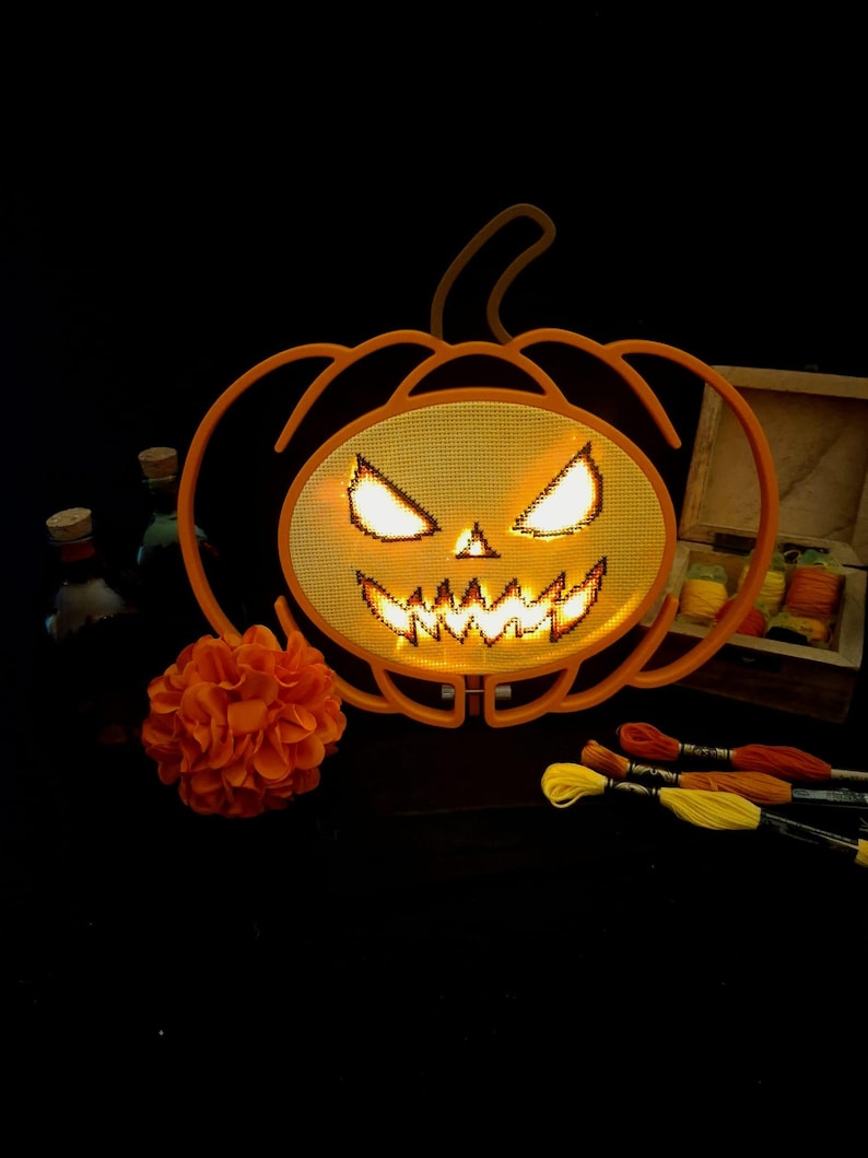 May include: An orange plastic pumpkin frame with a cross-stitch mesh center featuring a glowing jack-o'-lantern face. The design includes sharp, illuminated eyes and a jagged mouth. Nearby are an orange fabric flower and embroidery floss.