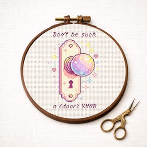 Könnte beinhalten: Ein Kreuzstich-Stickrahmen mit einem pixeligen Design eines Türknaufs mit einem bunten, kugelförmigen Griff. Der Text "Don't be such a (door) KNOB" ist über dem Türknauf gestickt. Eine goldene Schere liegt neben dem Rahmen.