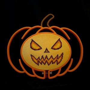 May include: A cross-stitch embroidery of a jack-o'-lantern face featuring glowing yellow and orange eyes and a jagged mouth, mounted within an orange plastic pumpkin-shaped frame against a solid black background.