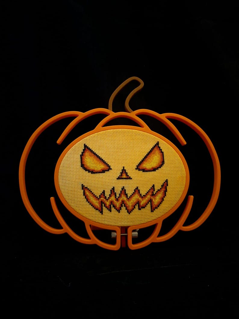 May include: A cross-stitch embroidery of a jack-o'-lantern face featuring glowing yellow and orange eyes and a jagged mouth, mounted within an orange plastic pumpkin-shaped frame against a solid black background.