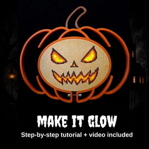 May include: An orange pumpkin-shaped embroidery hoop frame containing a cross-stitch design of a jack-o'-lantern face with glowing yellow and orange eyes and a jagged mouth. The text below reads MAKE IT GLOW Step-by-step tutorial + video included.
