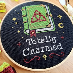May include: A circular wooden embroidery hoop containing black fabric with a cross-stitch design of a green book featuring a red triquetra symbol. Below the book, the text "Totally Charmed" is stitched in white, accented by red decorative swirls.