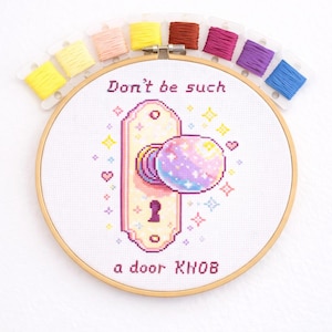 Pode incluir: Um bastidor de bordado em ponto cruz com o texto "Don't be such a door knob". O desenho apresenta uma maçaneta com um design celestial. Fio de bordar em várias cores é disposto acima do bastidor.