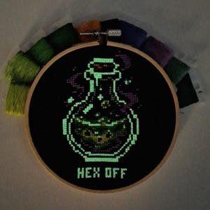 May include: A circular wooden embroidery hoop containing black fabric with a cross-stitched glowing green potion bottle and the text HEX OFF. The design features bubbling liquid inside the bottle and wisps of smoke, surrounded by colorful embroidery floss.