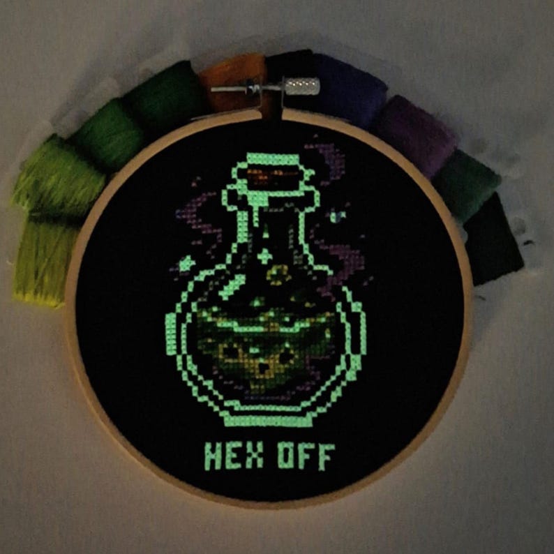 May include: A circular wooden embroidery hoop containing black fabric with a cross-stitched glowing green potion bottle and the text HEX OFF. The design features bubbling liquid inside the bottle and wisps of smoke, surrounded by colorful embroidery floss.