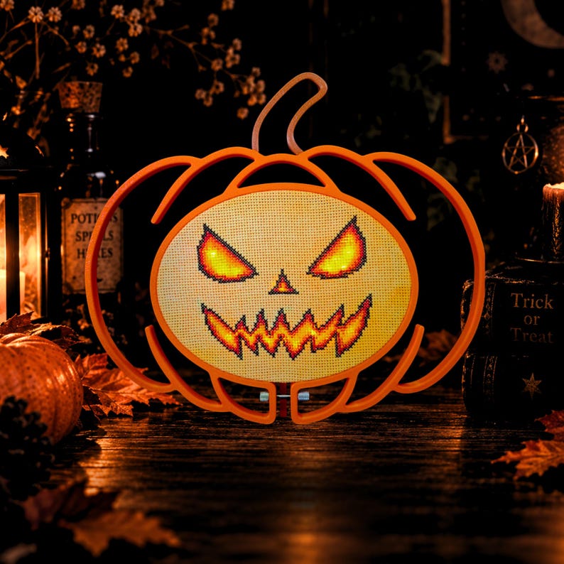 May include: A pumpkin-shaped light-up decoration featuring a cross-stitched face with glowing yellow and orange eyes and a jagged mouth. The frame is made of orange plastic, and the central panel displays a stitched jack-o'-lantern design.