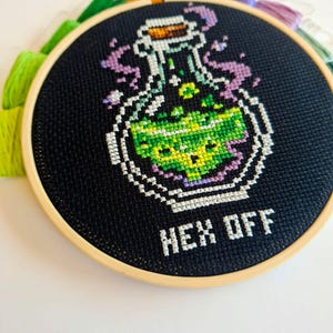 May include: A circular wooden embroidery hoop containing black fabric with a cross-stitch design of a glass potion bottle filled with bright green liquid. Purple smoke rises from the bottle, and the white text below reads HEX OFF.