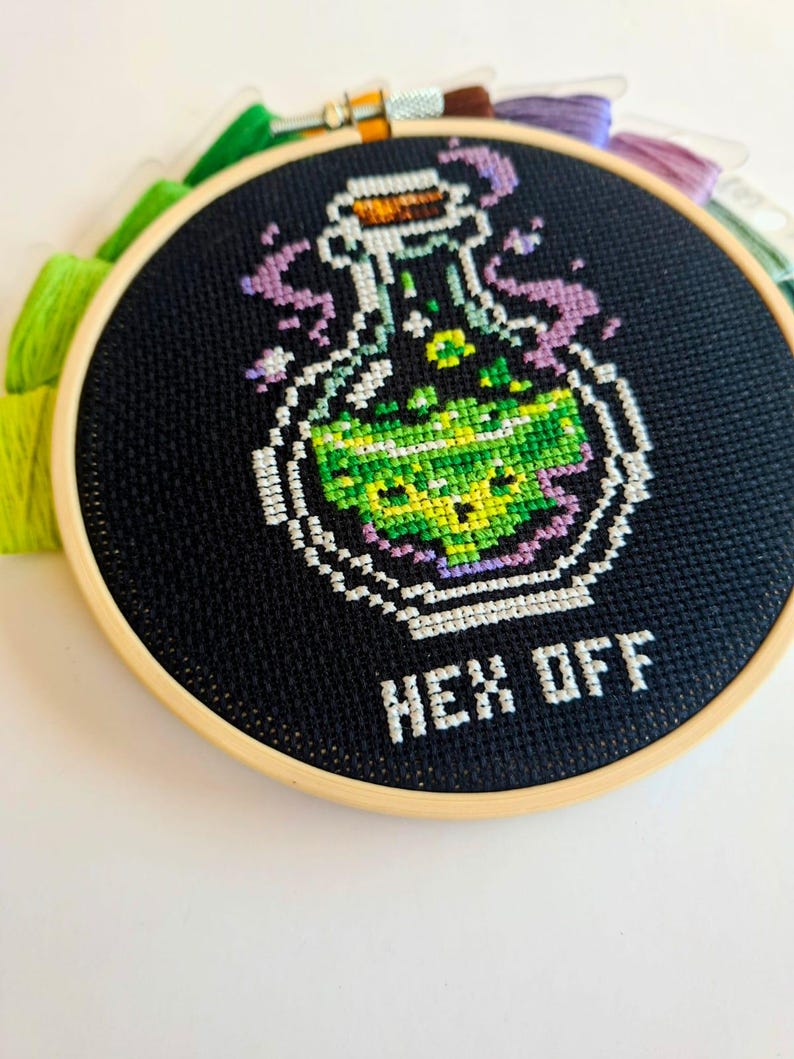 May include: A circular wooden embroidery hoop containing black fabric with a cross-stitch design of a glass potion bottle filled with bright green liquid. Purple smoke rises from the bottle, and the white text below reads HEX OFF.