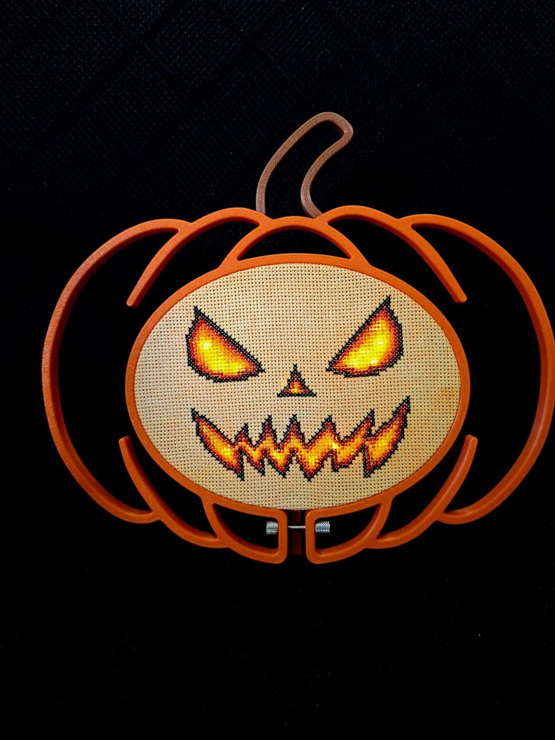 May include: A pumpkin-shaped orange embroidery hoop frame containing a cross-stitch design of a jack-o'-lantern face. The face features glowing yellow and orange eyes and a jagged mouth stitched onto light beige fabric, with a brown stem at the top.