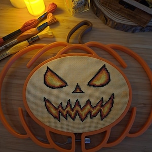 May include: A pumpkin-shaped embroidery hoop frame containing a cross-stitch pattern of a jack-o'-lantern face. The design features glowing yellow and orange eyes and a jagged, toothy mouth stitched onto light-colored fabric within the orange frame.