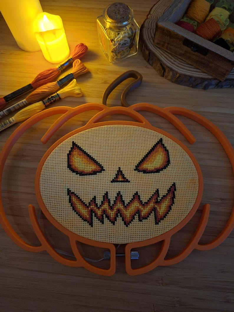 May include: A pumpkin-shaped embroidery hoop frame containing a cross-stitch pattern of a jack-o'-lantern face. The design features glowing yellow and orange eyes and a jagged, toothy mouth stitched onto light-colored fabric within the orange frame.