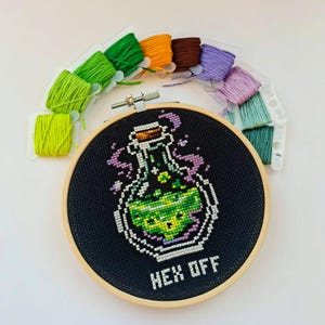 May include: A round wooden embroidery hoop containing black fabric with a cross-stitch design of a glass bottle filled with green liquid, purple smoke, and the text "HEX OFF" in white. Ten colorful embroidery floss bobbins are arranged above.