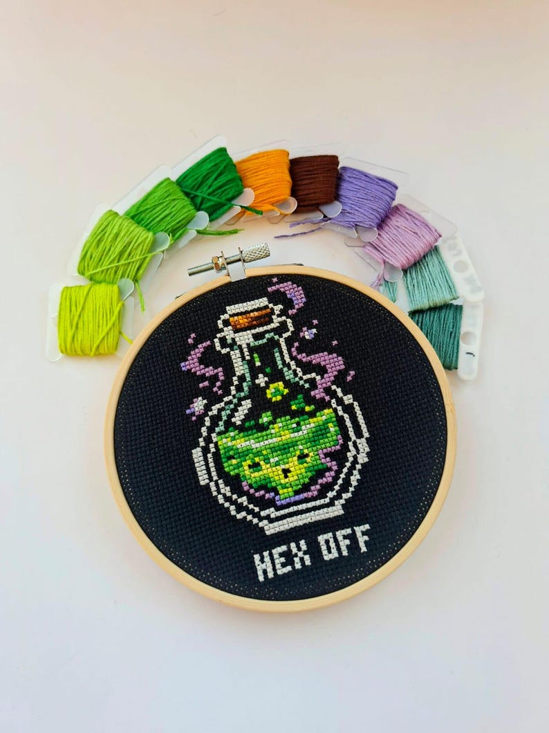 May include: A round wooden embroidery hoop containing black fabric with a cross-stitch design of a glass bottle filled with green liquid, purple smoke, and the text "HEX OFF" in white. Ten colorful embroidery floss bobbins are arranged above.