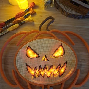 May include: A pumpkin-shaped embroidery hoop frame containing a cross-stitch fabric piece with a jack-o'-lantern face. The eyes and jagged mouth are illuminated from behind by warm yellow lights, creating a glowing effect on the fabric.