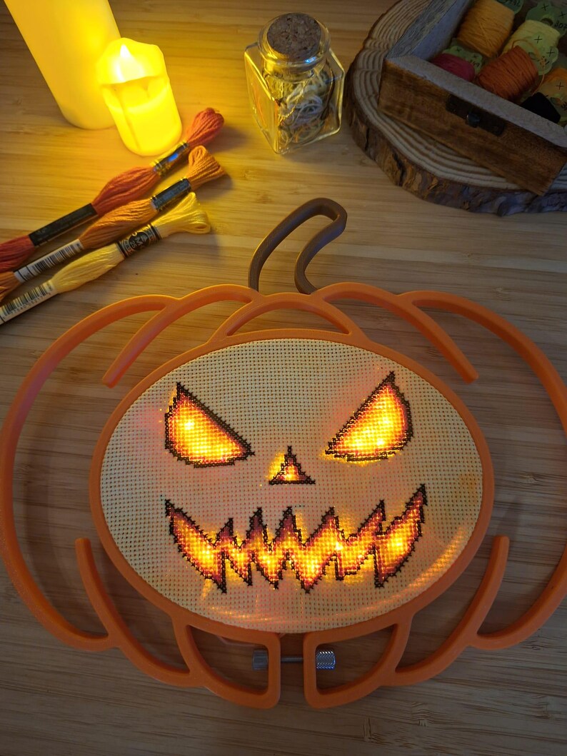May include: A pumpkin-shaped embroidery hoop frame containing a cross-stitch fabric piece with a jack-o'-lantern face. The eyes and jagged mouth are illuminated from behind by warm yellow lights, creating a glowing effect on the fabric.
