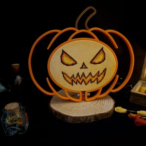 May include: An orange pumpkin-shaped frame containing a cross-stitch design of a jack-o'-lantern face with glowing yellow and orange eyes and a jagged mouth. The piece is mounted on a circular wooden base, with embroidery floss nearby.