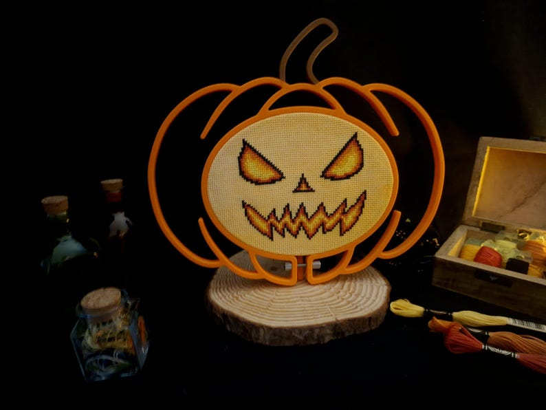 May include: An orange pumpkin-shaped frame containing a cross-stitch design of a jack-o'-lantern face with glowing yellow and orange eyes and a jagged mouth. The piece is mounted on a circular wooden base, with embroidery floss nearby.