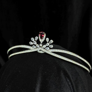 Op de afbeelding: Een zilverkleurige tiara met een dubbel bandontwerp. Het heeft een centrale, peervormige, rode edelsteen, omringd door kleinere, heldere edelstenen. De tiara staat tegen een zwarte achtergrond.