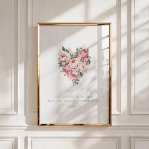 Könnte beinhalten: Gerahmter Druck mit goldenem Rahmen, der ein herzförmiges Blumenarrangement in Rosa und Weiß mit grünen Blättern zeigt. Der Druck enthält deutschen Text am unteren Rand. Das Kunstwerk wird an einer weißen Wand ausgestellt.