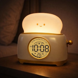 Op de afbeelding: Een nieuwigheid wekker in de vorm van een broodrooster, met een gloeiend, lachend toastvormig licht erbovenop. Het digitale display toont de tijd als 10:08 uur en de temperatuur als 26°C. De klok heeft een goudkleurige rand.