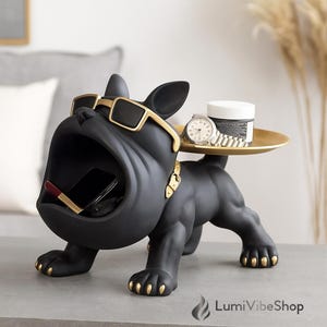 Può includere: Statuetta di bulldog nero con dettagli dorati, con occhiali da sole. Il bulldog ha la bocca aperta, che tiene un telefono e un rossetto. Un vassoio dorato sulla schiena contiene un orologio e un barattolo. Anche le zampe e il collare del cane sono dorati.