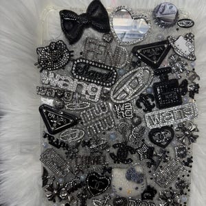 May include: A rectangular tablet case covered in a dense collage of silver and black decorative charms, including logos for Prada, Chanel, Fendi, Dior, and Miu Miu, alongside Hello Kitty figures, a black bow, a heart-shaped mirror, and rhinestones.