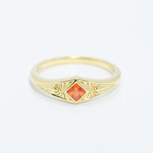 Op de afbeelding: Een gouden ring met een oranje vierkante edelsteen in een decoratieve, puntige zetting. De ring heeft een dunne band en staat tegen een witte achtergrond. De ring is een sieraad.