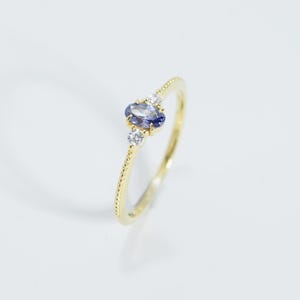 May include: A delicate gold ring featuring a central oval-cut, dark blue gemstone, flanked by two smaller, round, clear gemstones. The ring band has a textured, dotted design. The ring is set against a white background.