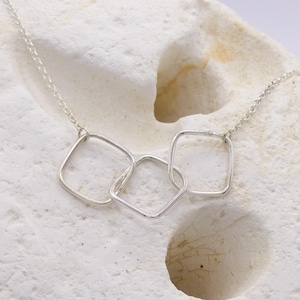 May include: A silver necklace featuring three interconnected square pendants. The delicate chain and pendants are made of a shiny, light-colored metal, likely silver. The necklace is displayed on a textured, off-white surface.