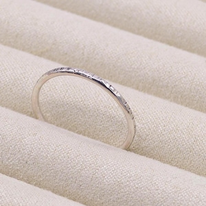 Könnte beinhalten: Ein zarter Silberring mit strukturierter Oberfläche. Der Ring ist dünn und hat ein schlichtes, elegantes Design. Der Ring wird auf einem hellbeigen Stoff präsentiert.