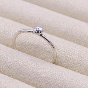 Puede incluir: Un anillo plateado con una pequeña bola texturizada en la parte superior. La banda del anillo es delgada y curvada, creando un diseño minimalista. El anillo se encuentra sobre una superficie beige claro y texturizada.