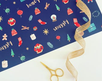 May include: A navy blue wrapping paper with a festive pattern of sweaters, socks, and gifts. The words "homey" and "snuggly" are written on the paper. A gold pair of scissors and a burlap ribbon are also present.