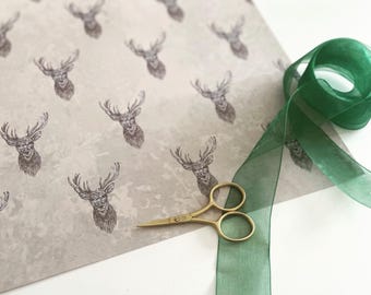 May include: A roll of wrapping paper with a repeating pattern of deer heads in brown ink on a light gray background. A green ribbon and gold scissors are also in the image.