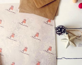 May include: Wrapping paper featuring a repeating pattern of watercolour robins on branches. Two brown envelopes, a pine cone ornament, and a silver star decoration are also present. The scene is set on a white wooden surface.