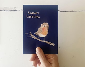 May include: A navy blue greeting card featuring a watercolor illustration of a robin perched on a branch. The text "Season's tweetings" is written in a gold script. The bird has a red breast and brown feathers.