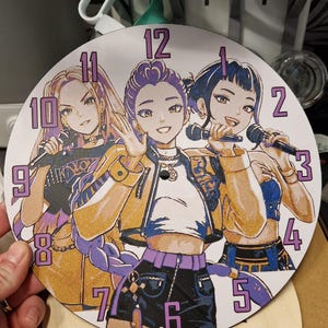 Può includere: Orologio rotondo con un'illustrazione di tre donne in stile anime che tengono microfoni. Il quadrante dell'orologio ha numeri viola e uno sfondo dorato scintillante. Le lancette dell'orologio sono rosse e dorate. Un oggetto decorativo.