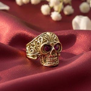 May include: A gold-colored skull ring with red gemstone eyes. The ring is intricately detailed with filigree patterns and sits on a deep red, silky fabric. White flower blossoms and petals are in the background.
