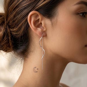 May include: A long, silver-toned threader earring featuring a delicate, wavy, S-shaped metal wire design that hangs vertically from the earlobe, ending in a thin, straight chain segment that extends downwards along the side of the neck.