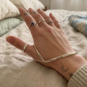 May include: A silver hand bracelet featuring a V-shaped design that connects the wrist to the thumb. The hand is adorned with three silver rings: one plain band on the thumb, a wave-shaped ring with blue inlay on the index finger, and a chain-link ring.