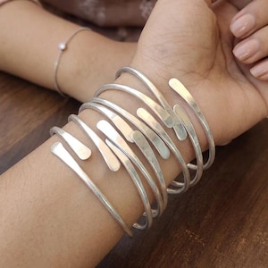 May include: A collection of silver-toned metal bracelets with a unique, open design. The bracelets are stacked on a wrist, showcasing their curved and elongated shapes. A delicate silver bracelet with a small pearl is also visible.