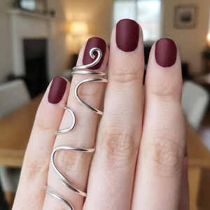 May include: Close-up of a hand with burgundy nail polish and silver wire rings. The rings have a unique, flowing design that wraps around the fingers. The nail polish is a matte finish.