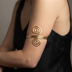 May include: A gold-colored metal arm cuff featuring a triple-band design that connects two symmetrical spiral motifs at the front. The piece is constructed from smooth, polished wire and is worn around the upper arm, showcasing a continuous circular form.
