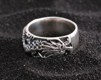 Può includere: Un anello d'argento con un design dettagliato di drago che avvolge la fascia. L'anello è realizzato in metallo lucido e poggia su una superficie scura e strutturata. Le squame e i dettagli del drago sono intagliati in modo intricato.