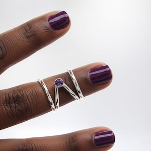 Puede incluir: Un anillo de plata con una pequeña gema púrpura, diseñado para envolver un dedo. El anillo se muestra en una mano de piel oscura con esmalte de uñas púrpura.