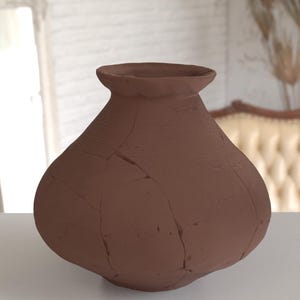 May include: A brown, handcrafted ceramic vase with a wide, rounded base and a narrow neck. The vase has a textured surface with visible cracks, adding to its rustic aesthetic. The vase is approximately 10 inches tall.