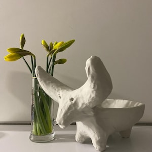 Pode incluir: Uma escultura de cerâmica branca de um animal com um corpo em forma de tigela e três pernas. Narcisos amarelos em um vaso de vidro transparente estão à esquerda da escultura. O fundo é uma parede branca lisa.
