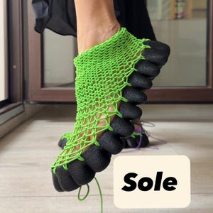 Könnte beinhalten: Ein limettengrünes Obermaterial aus Kordel im Makramee-Stil, befestigt an einer dicken, schwarzen, strukturierten und segmentierten Gummisohle. Das Schuhwerk hat eine netzartige Konstruktion, die den Fuß umschließt, mit dem Wort Sole auf einem Etikett.