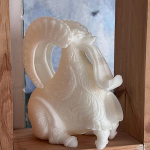 Puede incluir: Una figura de carnero impresa en 3D de color blanco translúcido en posición sentada. La escultura presenta cuernos curvos detallados, patrones de pelaje texturizados en el cuerpo y una abertura cilíndrica hueca en la parte inferior frontal.