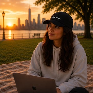 May include: A black baseball cap with the text "vibe coder" in white, worn by a person. The person is wearing a light gray hoodie and sitting on a blanket with a laptop. The background features a city skyline at sunset.